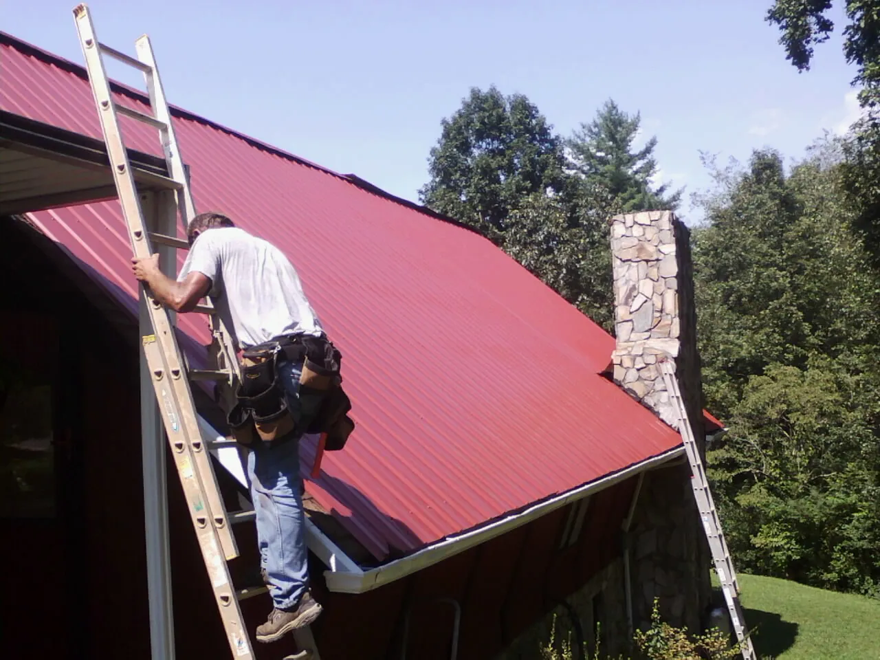 Roofer on a ladder beside a red metal roof and stone chimney — Soffit & Fascia Repair by Roofing Brothers in Grapevine